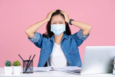 Genç, yorgun Asyalı iş kadını gündelik giysiler içinde ve maske takarak 19 numaralı ofisteki bilgisayarla masa başında oturmasını önlüyor. Pembe arka plan stüdyosunda elleri izole bir şekilde kafasına dokundu..
