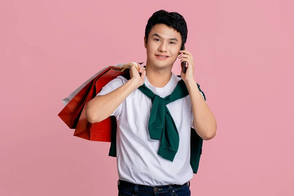 ピンクの背景に隔離されたスマートフォンで話すショッピングバッグを持つ若い男 — ストック写真