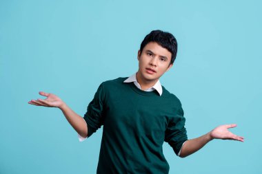 Açık mavi arka planda yakışıklı bir Asyalı. Kolları ve elleri havada, budala ve kafası karışık bir ifade takındı.