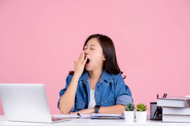 Sıradan gömlekler giyen yorgun Asyalı kadın beyaz ofis masasında çalışıyor. Pembe arka planda tek başına esniyor.