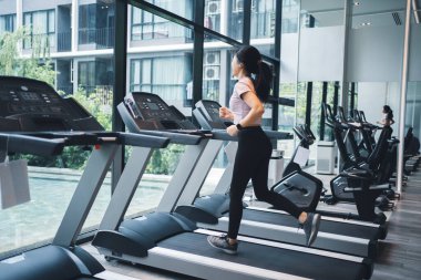 Spor salonundaki büyük bir pencerenin yanında koşu bandında koşan Asyalı bir kadın. Sağlıklı yaşam tarzı kavramı.