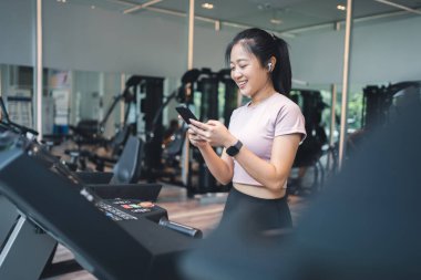 Koşu bandında çalışan kadın, kablosuz kulaklık takıyor ve akıllı saat cep telefonunu kontrol ediyor..