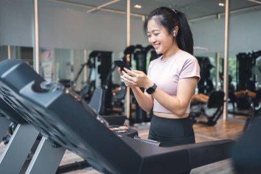Genç bir kadın koşu bandında egzersiz yapıyor ve spor salonunda akıllı telefon kullanıyor..