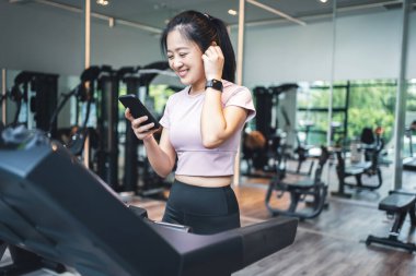 Koşu bandında egzersiz yapan ve spor salonunda fotokopi aletiyle müzik dinleyen genç bir kadın.