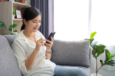 Beautiful Asian woman relaxing on comfortable sofa, enjoying web surfing information in internet or using mobile software application at home.