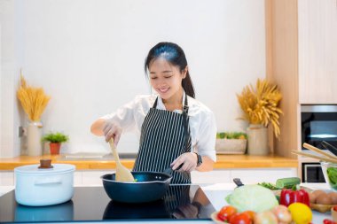 Asian woman standing by the stove in the kitchen, she is cooking and smelling the aroma of her food in the pot.
