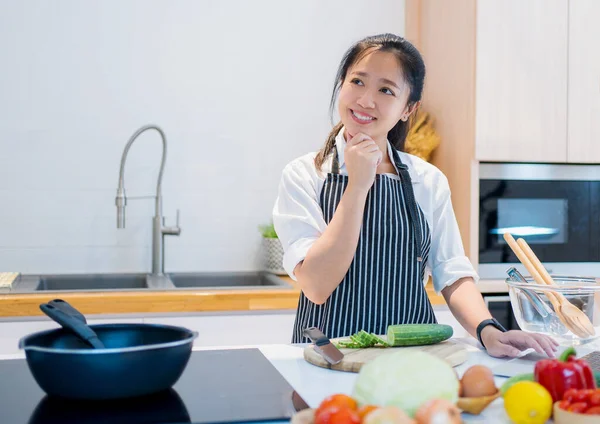 Young happy asian smiling woman is thinking of recipes in the kitchen. Healthy lifestyle concept. Girl cooking at home prepare food.