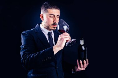 Siyah giyinmiş zarif şarap garsonu kırmızı şarap kadehi gibi kokar elinde yatay çekimde koyu arka planda bir şişe tutar.