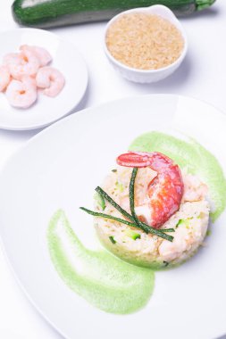Beyaz arka planda şef tarafından pişirilmiş karides ve kabaklı risotto ve arkasında malzemeler ve çok parlak ışık dikey çekim