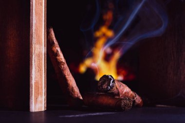 Purolar, yanan bir puroyla karanlık bir yüzeyde, ahşap çerçevede duman çıkarır ve arka planda, lüks bir atmosferde şöminenin alevleri vardır.