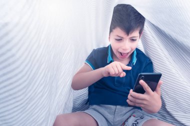 Akıllı telefon bağımlısı beyaz çizgili çarşafların altında elinde çok heyecanlı bir ifadeyle akıllı telefon tutan bir çocuk.