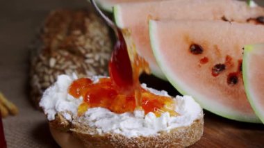 Bir dilim beyaz peynirli ekmeğe kaşıkla reçel sür. Yakın plan, hafif doğal kamera titremesi..