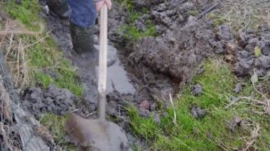Lastik botlu bir adam su akıntısı için bir kanalı kürekle temizliyor. Kirli ve zor fiziksel çalışma.