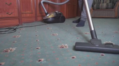 Adam evi temizliyor. Halıyı süpürüyor. Çerçevede sadece bacakları ve elektrikli süpürge görünüyor. Alt açı.