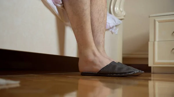 Men\'s feet shod in slippers near the bed. With one hand, the man holds on to his leg.