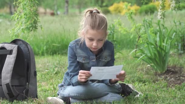 小学生の女の子が芝生の上の公園に座り 注意深く読む 現代の子供たちの日常生活 中学校での教育 — ストック動画