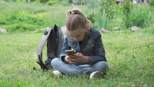 9歳の少女が芝生の上の公園に座り スマートフォンにテキストメッセージを入力します 彼女はスマートフォンを使ってソーシャルネットワークを閲覧しています — ストック動画