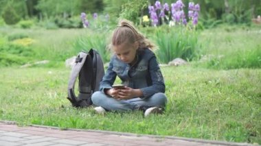 Bir kız, bir ilkokul öğrencisi, parktaki yeşil çimlerin üzerinde oturuyor. Akıllı telefonuna göz atmaya dalmış, okul çantası yakınlarda..