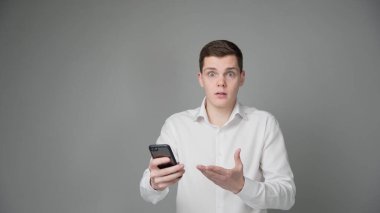 Şaşırmış bir genç adam akıllı telefonunun ekranına bakıyor. Gri bir arka planda. Yüksek kalite fotoğraf