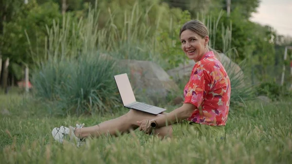Genç bir kadın parkta çimlerin üzerinde oturuyor ve önünde dizüstü bilgisayarını tutuyor. Kameraya bakarken gülümsüyor..