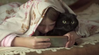 Genç kız kedisiyle battaniyenin altında uzanıyor ve akıllı telefonuyla oyun oynuyor. 4k video.