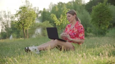 Orta yaşlı bir kadın parkta çimlerin üzerinde oturur. Bir dizüstü bilgisayardaki video bağlantısını kullanarak bir konuşma yapıyor. Yüksek kalite 4k görüntü