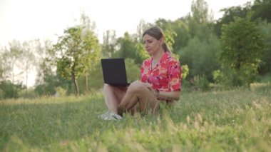 Orta yaşlı bir kadın parkta çimlerin üzerinde oturur. Bir dizüstü bilgisayardaki video bağlantısını kullanarak bir konuşma yapıyor. Yüksek kalite 4k görüntü
