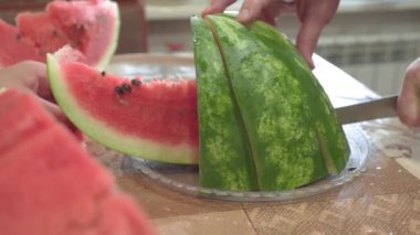 Erkeklerin elleri olgun kırmızı karpuzu bıçakla parçalara ayırır ve tabağa serer. Yakın plan..