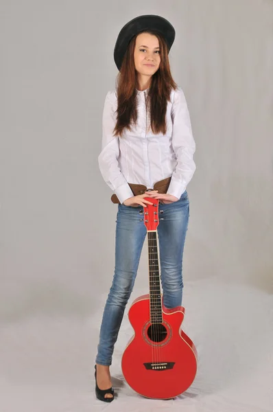 La ragazza con un sorriso imbarazzato in un cappello nero, camicia bianca e jeans si basa sulla chitarra rossa — Foto Stock