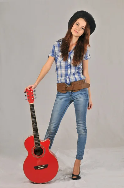 Cunningly smiling girl in a black hat, white shirt, jeans and cowboy belt relies on red guitar — Stock Photo, Image