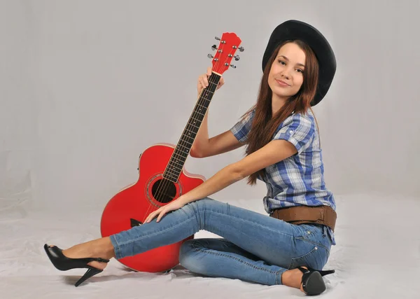 Chica joven sentada en una blusa con mangas cortas, su mano derecha en el cuello de la guitarra roja, y la izquierda se basa en un estirado hacia adelante con la pierna derecha — Foto de Stock