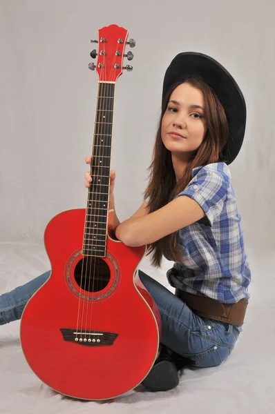 A smiling girl in a black hat, a plaid shirt, jeans and cowboy belt sitting hugs red guitar — Stock Photo, Image
