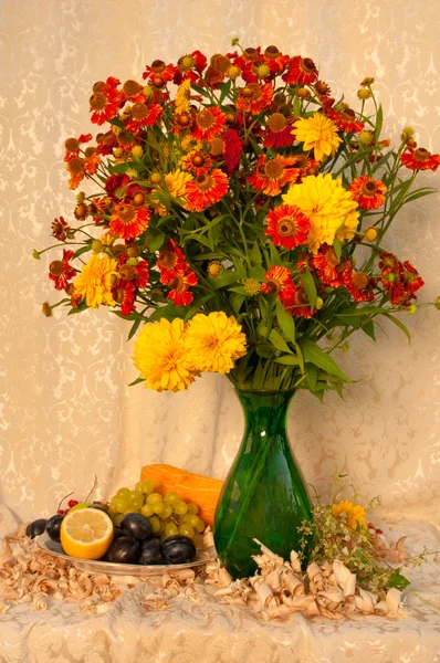 Натюрморт с ярко-желтыми и оранжевыми цветами в зеленой вазе с фруктами — стоковое фото