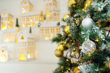 Oyuncak ve çelenkle süslenmiş Noel ağacı dalları beyaz bir merdivenin arka planında parlayan pencereli mukavva evlerle alay ediliyor. Noel ve yeni yıl arkaplanı