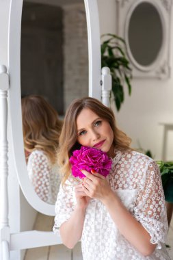 Beyaz elbiseli bir kadın elinde şakayık tomurcuğuyla kameraya bakıyor. Güzel sarışın kadın portresi. Randevu için sürpriz bir buket çiçek, anneler günü ya da sevgililer günü. Metnin için yer.