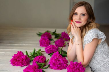 Kısa elbiseli genç bir kadın ahşap zeminde, pembe şakayık çiçeklerinin yanında oturuyor. Mesajın için yer aç. Güzellik ve gençlik kavramı. Çiçekçi ve tasarımcı