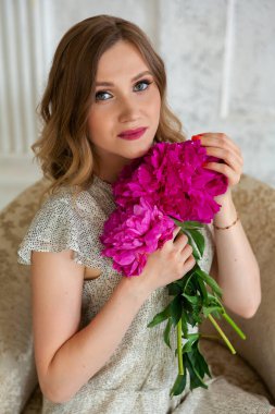 Beyaz elbiseli kadın stüdyodaki beyaz odada bir koltukta oturuyor elinde şakayık tutuyor ve kameraya bakıyor. Sakin ol.