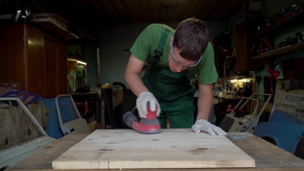 Timmerman in beschermende bril is gelijk aan polijst houten plaat met baanschuurmachine in werkplaats — Stockvideo