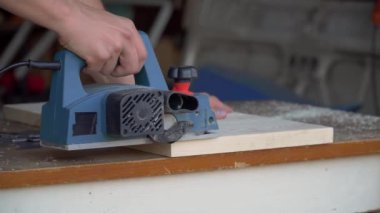 Marangoz, atölyedeki ahşap tahtada elektrikli planörle çalışıyor. Zanaatkar masayı yapar
