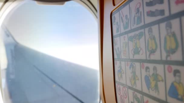 Instruções de segurança fotos no assento do avião e visão turva da janela na asa plana e céu - 4K, Editorial, Handheld — Vídeo de Stock