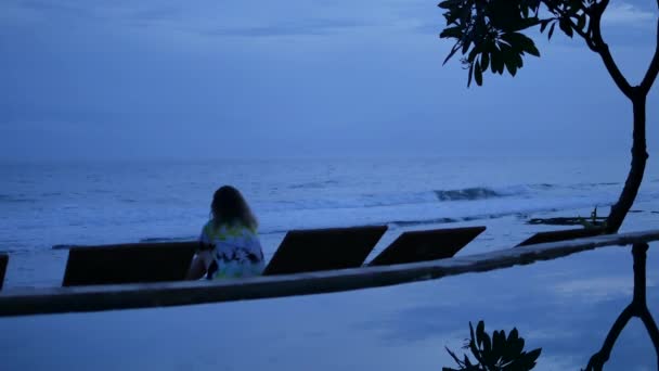 Mulher senta-se na praia perto da piscina e olha para as ondas oceânicas em uma hora azul em Amed, Bali - 4K, Áudio, Handheld — Vídeo de Stock