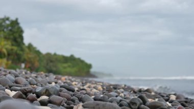 Volkanik çakıl taşlı plaj sabah Amed, Bali 'de arka planda yeşil ağaçları ve deniz dalgalarını bulanıklaştırdı - 4K, Audio, Medium shot