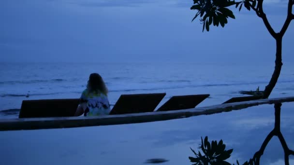 Woman at the dusk sits on the beach, straightens her hair and looks at oceanic waves in Amed, Bali - 4K — Vídeo de Stock