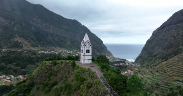 Pequeña iglesia rodeada de acantilados y montañas y un pueblo en un paisaje de naturaleza verde. En Madeira Portugal. — Vídeo de stock