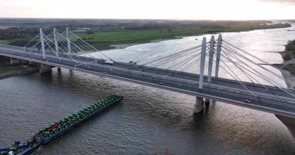 Ewijk, 9th of april 2022, The Netherlands. Commercial transportation ships passing under a suspension bridge with highway and traffic over it. Aerial drone view Sunset — ストック動画