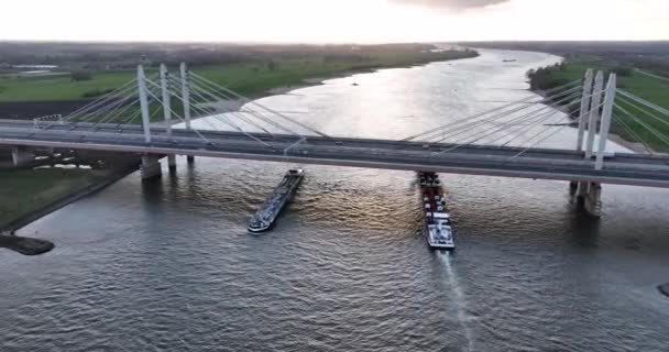 Ewijk, 9 kwietnia 2022, Holandia. Komercyjne statki transportowe przejeżdżające pod mostem wiszącym z autostradą i ruchem nad nim. Widok z lotu ptaka Zachód słońca — Wideo stockowe