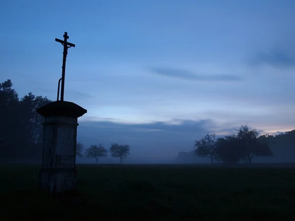 Katholisches Kruzifix auf der Wiese im ersten Abendnebel, alte Bäume am Horizont, blaue Wolken freier Nachthimmel. — Stockfoto