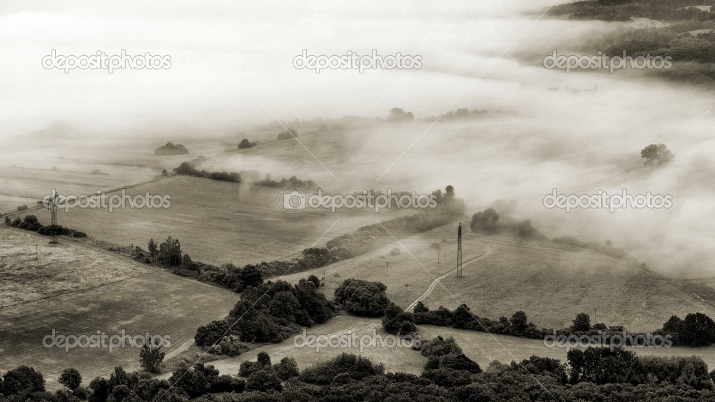 View into foggy countryside with bushes, forests, villages, levels of ponds within early morning sunrise.