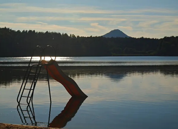 Abandoned old children slide stays in pond at the end of summer , reflection in blue water level, forest at horizon. — Stock Photo, Image