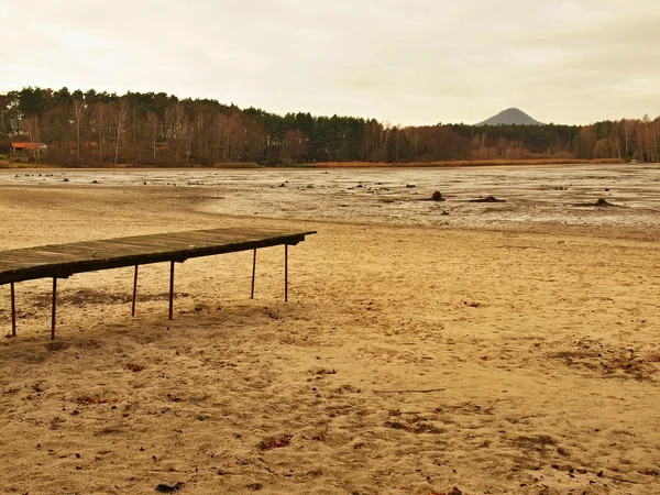 Abandoned beach at empty pond, old wooden mole above dirty beach sand. Autumn melancholic atmosphere. — Stock Photo, Image